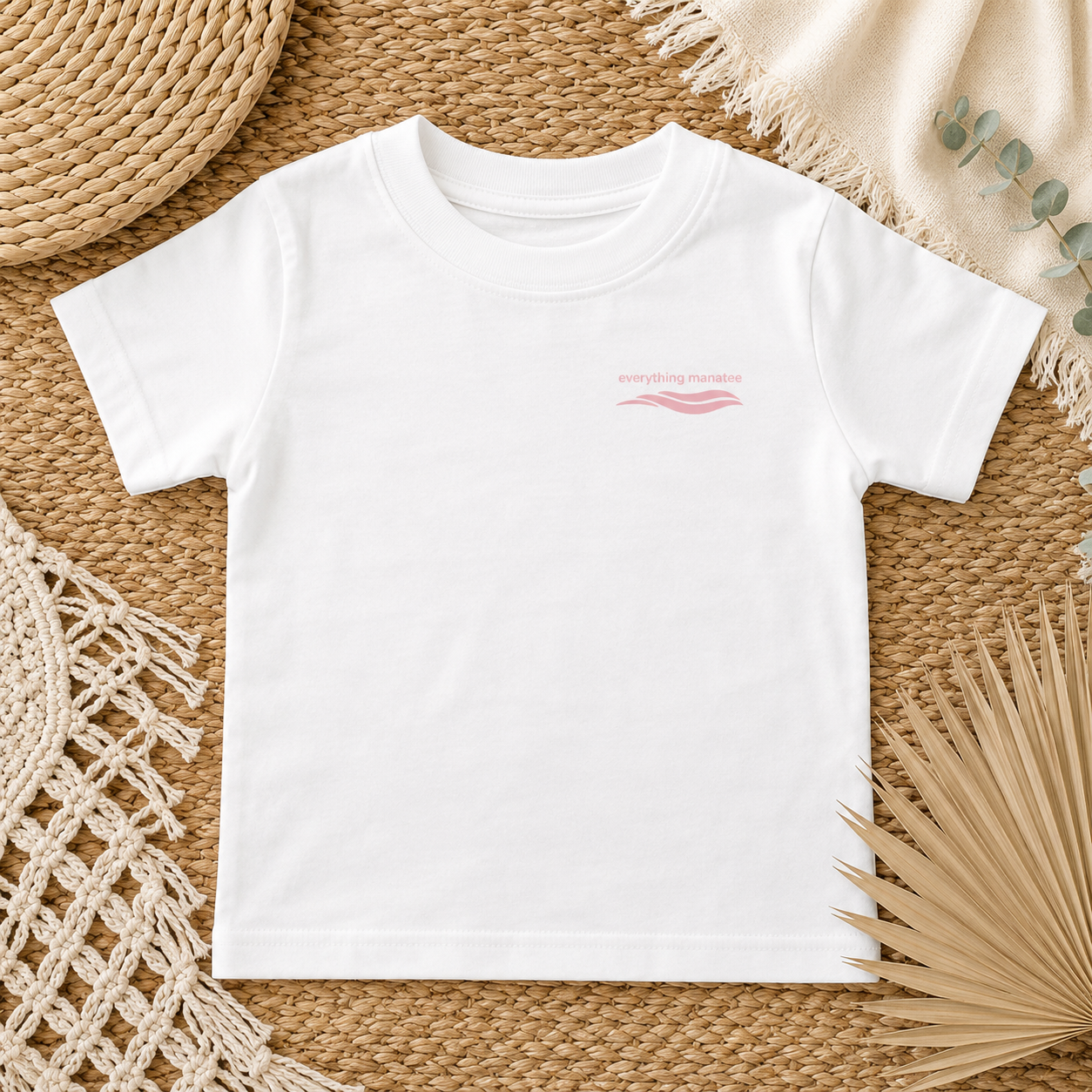 A white short-sleeve crewneck t-shirt with a small pink graphic on the left chest, laid flat on a woven straw mat with neutral decorative props.