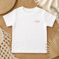 A white short-sleeve crewneck t-shirt with a small pink graphic on the left chest, laid flat on a woven straw mat with neutral decorative props.