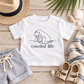 A white short-sleeve baby T-shirt laid flat on a rustic wood surface, featuring a black line drawing of a manatee and the words 'coastal life.' Surrounding props include a straw hat, blue-and-white striped shorts, beige sandals, shells, palm leaves, and a striped knit blanket.