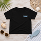 A black short-sleeve t-shirt with a small light blue 'everything mariner' wave logo on the left chest, presented in a flat-lay arrangement with seashells, sunglasses, a palm leaf, and a striped towel on a whitewashed wood background.