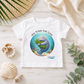 A white short-sleeve baby t-shirt laid flat on a light wood surface, surrounded by beach-themed props including shells, a woven mat, palm leaves, and a cream fabric. The shirt features a circular graphic of a manatee holding green seaweed under the text 'HUG WHAT YOU LOVE'.