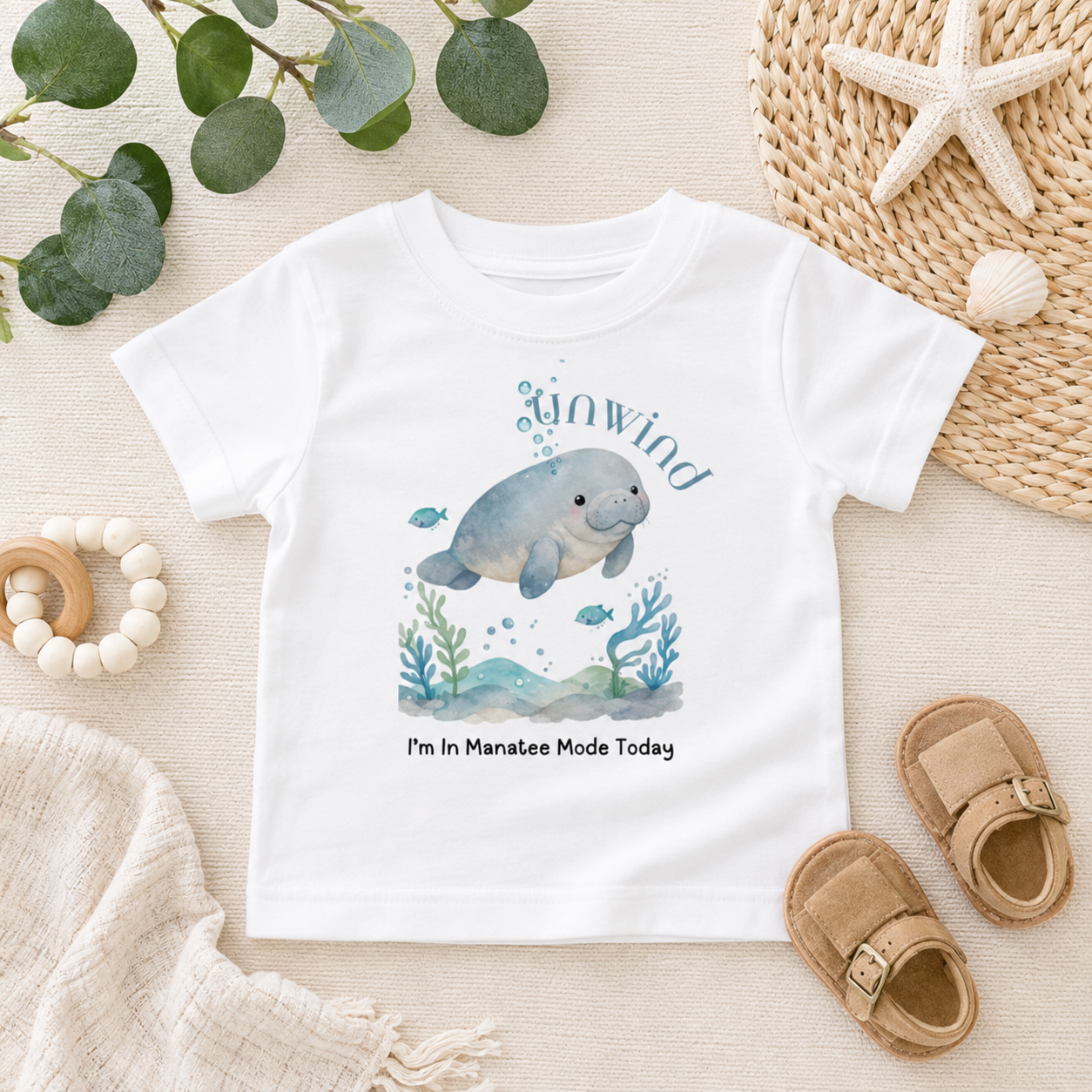 A white short-sleeve baby T-shirt laid flat on a light fabric surface, featuring a watercolor manatee illustration with the words 'Unwind' and 'I'm In Manatee Mode Today.' The shirt is styled with eucalyptus leaves, a woven hat, a starfish, baby sandals, and a wooden bead ring.