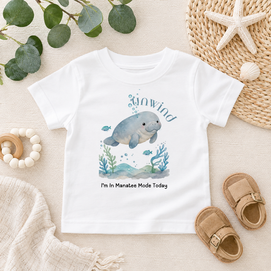A white short-sleeve baby T-shirt laid flat on a light fabric surface, featuring a watercolor manatee illustration with the words 'Unwind' and 'I'm In Manatee Mode Today.' The shirt is styled with eucalyptus leaves, a woven hat, a starfish, baby sandals, and a wooden bead ring.