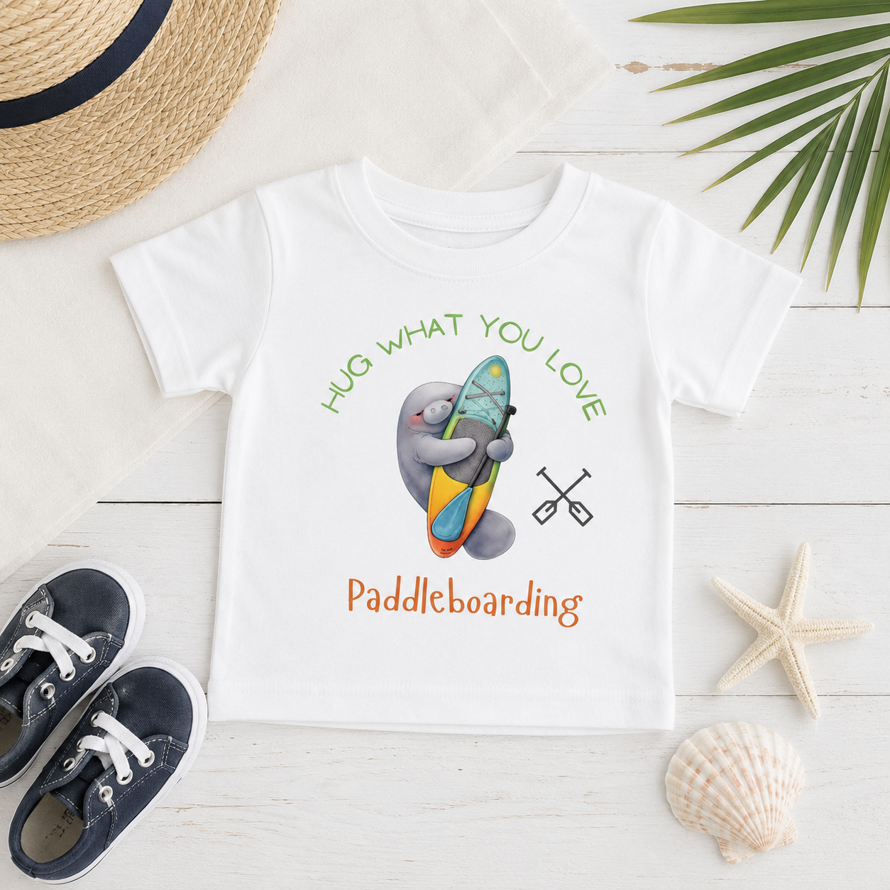 A white short-sleeve kids' T-shirt laid flat on a white wood surface, featuring a cartoon gray animal hugging a colorful paddleboard with the text 'HUG WHAT YOU LOVE' above and 'Paddleboarding' below. The shirt is styled with a straw hat, navy sneakers, a starfish, a seashell, and palm leaves.