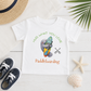 A white short-sleeve kids' T-shirt laid flat on a white wood surface, featuring a cartoon gray animal hugging a colorful paddleboard with the text 'HUG WHAT YOU LOVE' above and 'Paddleboarding' below. The shirt is styled with a straw hat, navy sneakers, a starfish, a seashell, and palm leaves.