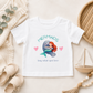 A white toddler t-shirt featuring a cute graphic of a mermaid hugging a blue whale, with the text 'MERMAIDS' and 'hug what you love'. The shirt is displayed in a styled flat-lay setting with a beige blanket, baby sandals, a wooden toy boat, and a wicker basket.
