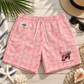 Pink patterned swim shorts with white drawstrings, laid flat on a light wood surface with tropical and beach accessories around them. The shorts have a tonal leaf pattern, a small white brand patch on the left hip, and a brown polar bear graphic with text on the lower right leg.