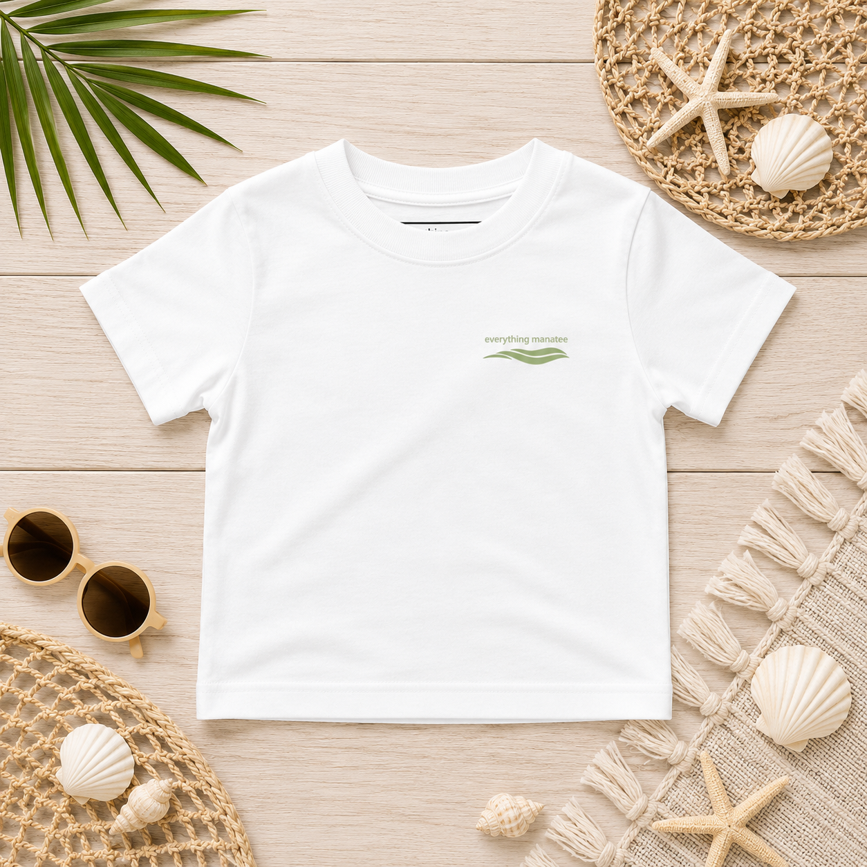 A white short-sleeve t-shirt with a small green 'everything natural' logo on the left chest, styled in a flat lay on a light wood background with beach-themed props.