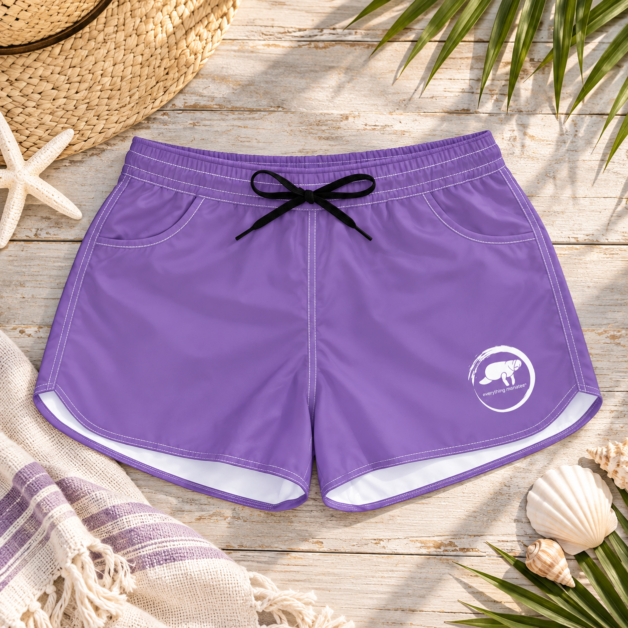 Purple swim trunks with a black drawstring and a white logo on the lower left leg, laid flat on a rustic wooden surface with beach-themed props including a straw hat, starfish, seashells, and palm leaves.