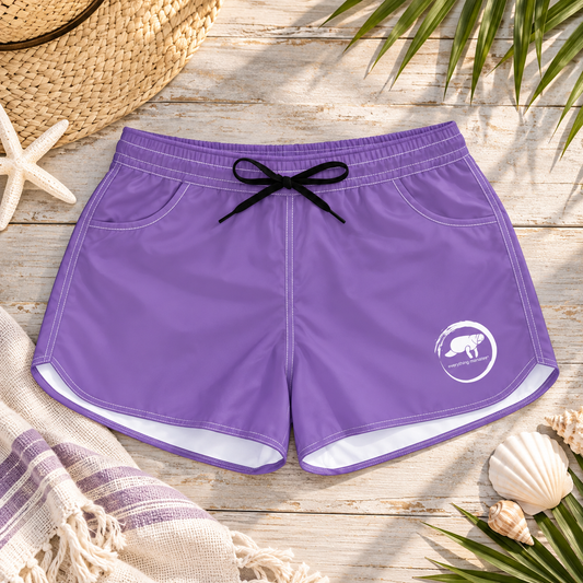 Purple swim trunks with a black drawstring and a white logo on the lower left leg, laid flat on a rustic wooden surface with beach-themed props including a straw hat, starfish, seashells, and palm leaves.