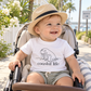 A baby sitting in a stroller outdoors wearing a white t-shirt with a black graphic of a manatee and the text 'coastal life', along with a straw hat and light gray shorts.