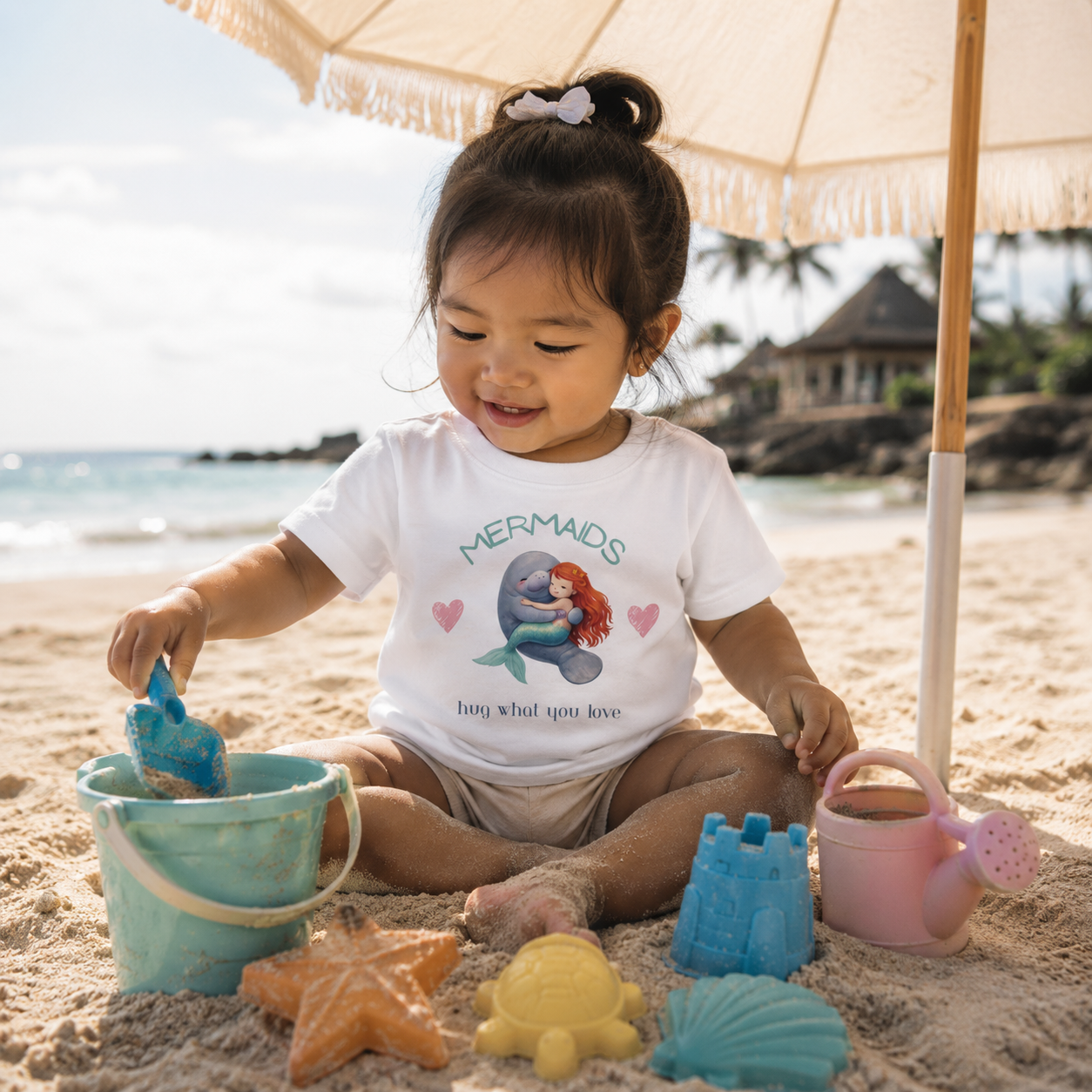 A toddler girl sitting on a sandy beach under a beige umbrella, wearing a white graphic t-shirt with a mermaid design. She is surrounded by colorful plastic beach toys including a blue bucket, starfish, seashell, and watering can.