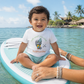 A smiling toddler sits cross-legged on a turquoise and white paddleboard on blue water, wearing a white graphic T-shirt that reads 'HUG WHAT YOU LOVE' and 'Paddleboarding' with a heart-and-surfboard illustration. A real outdoor beach environment with palm trees and water is visible in the background, so this is Lifestyle rather than Product/Packaging.