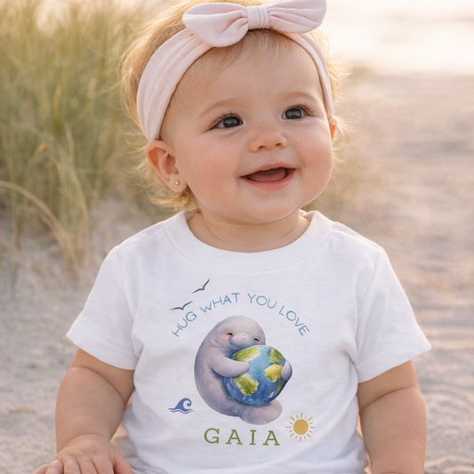 Baby sitting on sand wearing a white t-shirt with a graphic design and text.
