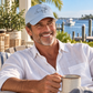 A smiling man wearing a light blue baseball cap with a simple line drawing of a dog, seated outdoors by a waterfront. He is wearing a white button-down shirt and holding a mug.