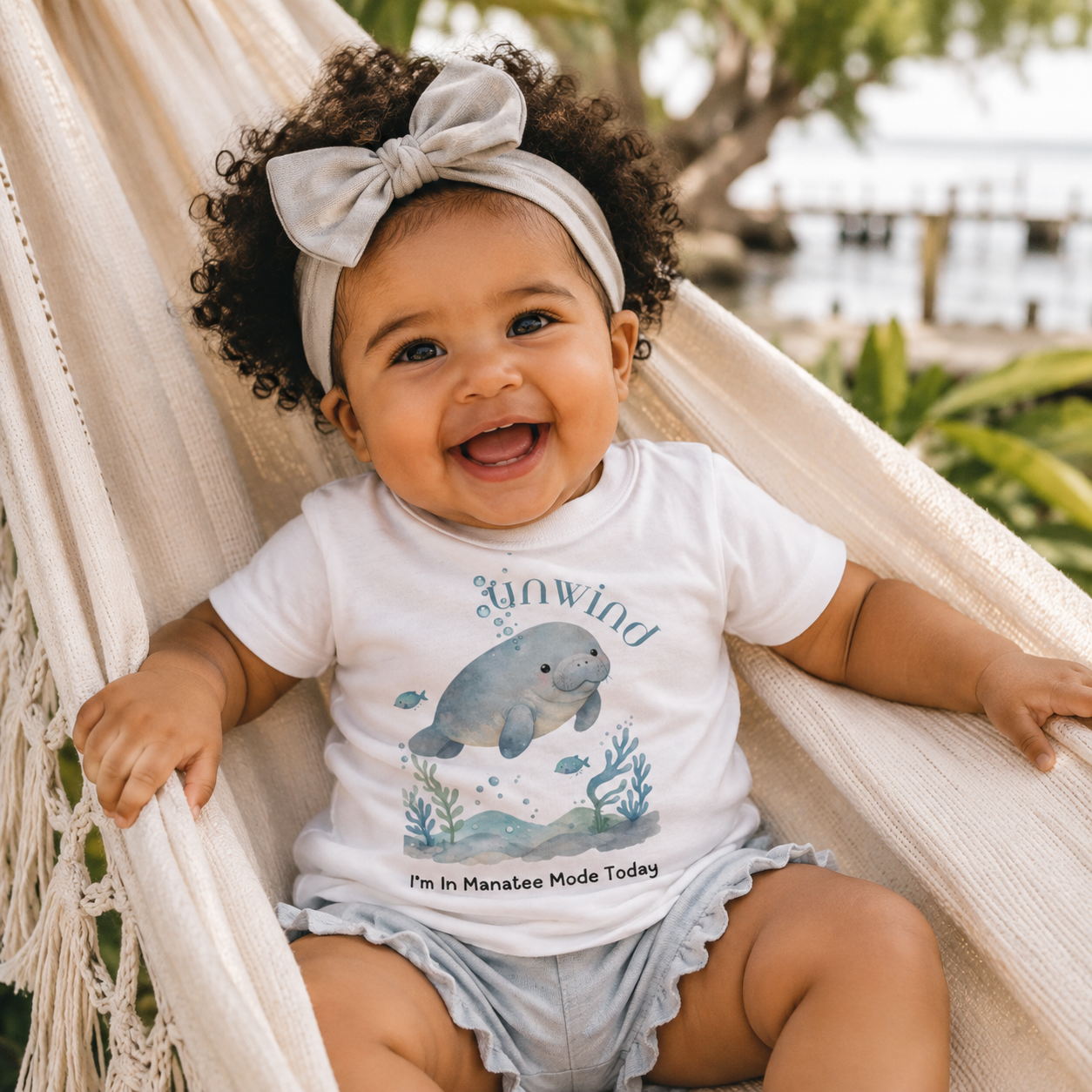 A smiling baby sitting in a cream hammock outdoors, wearing a white short-sleeve graphic T-shirt with a manatee illustration and the words 'Sunwind' and 'I'm In Manatee Mode Today,' along with light denim shorts and a matching bow headband.