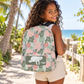 A smiling young girl with curly hair is shown from behind wearing a tropical print backpack on a sunny beach path with palm trees and the ocean in the background.