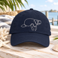 A navy blue dad hat with a white line-art embroidered manatee on the front, styled on a wooden table with beach-themed props like a starfish, seashell, and a striped towel, with a blurred waterfront background.