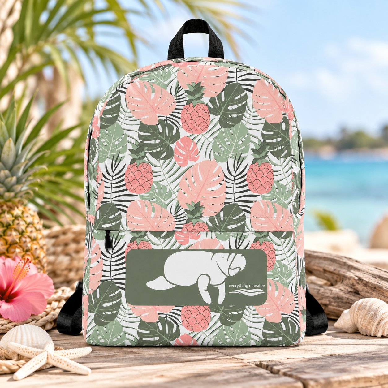 A backpack with a tropical monstera and pineapple print in pink, green, and black, featuring a front pocket with a manatee graphic and the text 'The Manatee Company'. The backpack is displayed on a wooden beach table with tropical props like a pineapple, hibiscus flower, seashells, and driftwood, with a blurred beach background.