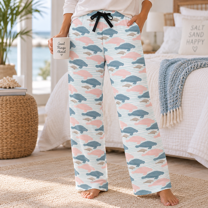 A person wearing light blue wide-leg pajama pants with a pink and blue cloud pattern, standing barefoot in a bright bedroom setting and holding a white mug.