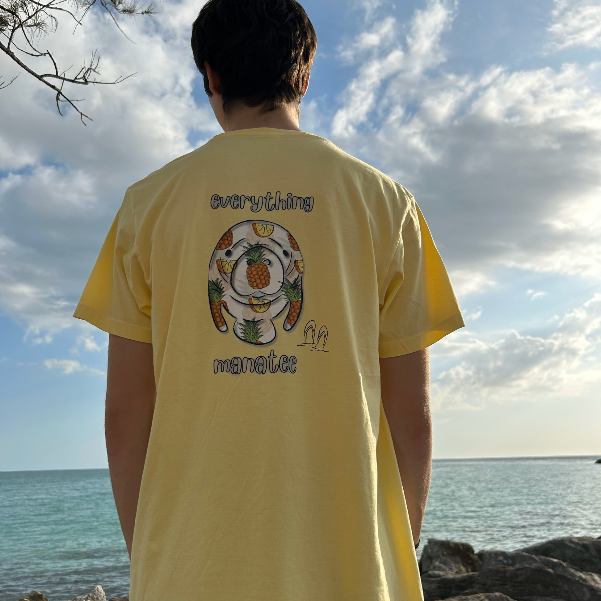 Person wearing Pineapples & Paradise Manatee T-Shirt in banana yellow, styled with ocean view in the background