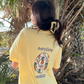 Person wearing Pineapples & Paradise Manatee T-Shirt in banana yellow, styled with palm trees in the background