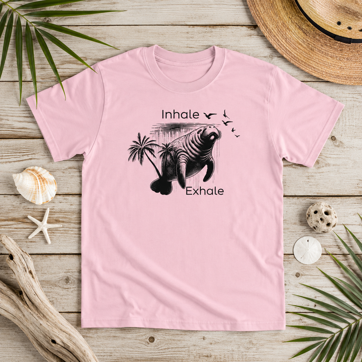A light pink short-sleeve t-shirt with a black graphic of a manatee and the words 'Inhale' and 'Exhale' laid flat on a rustic wooden surface surrounded by beach-themed props like a straw hat, starfish, shells, driftwood, and palm leaves.