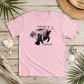 A light pink short-sleeve t-shirt with a black graphic of a manatee and the words 'Inhale' and 'Exhale' laid flat on a rustic wooden surface surrounded by beach-themed props like a straw hat, starfish, shells, driftwood, and palm leaves.