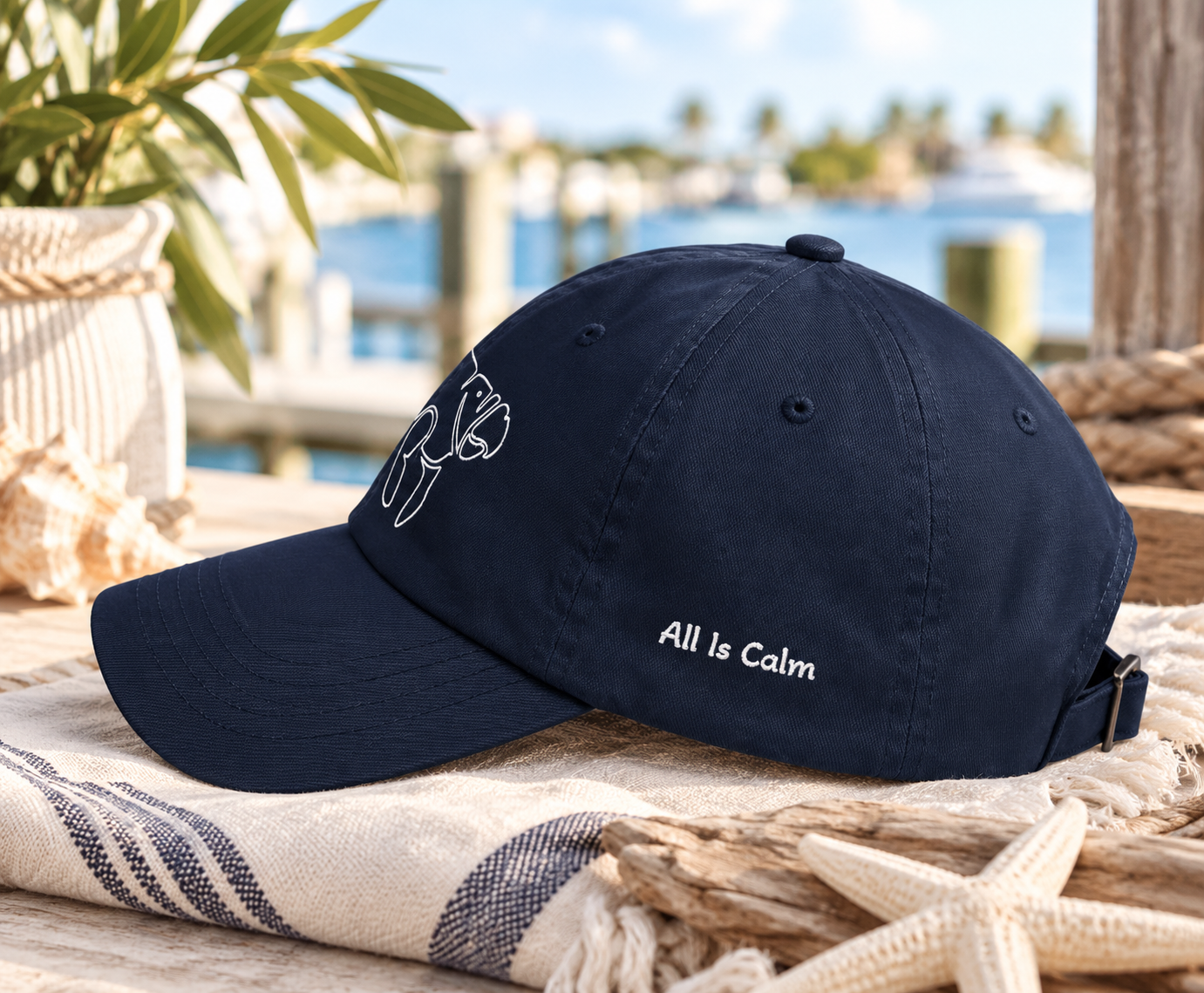 A navy blue baseball cap with white embroidered text reading 'All Is Calm' and a white line-art graphic on the front, styled on a beach-themed surface with rope, a towel, and a blurred waterfront background.